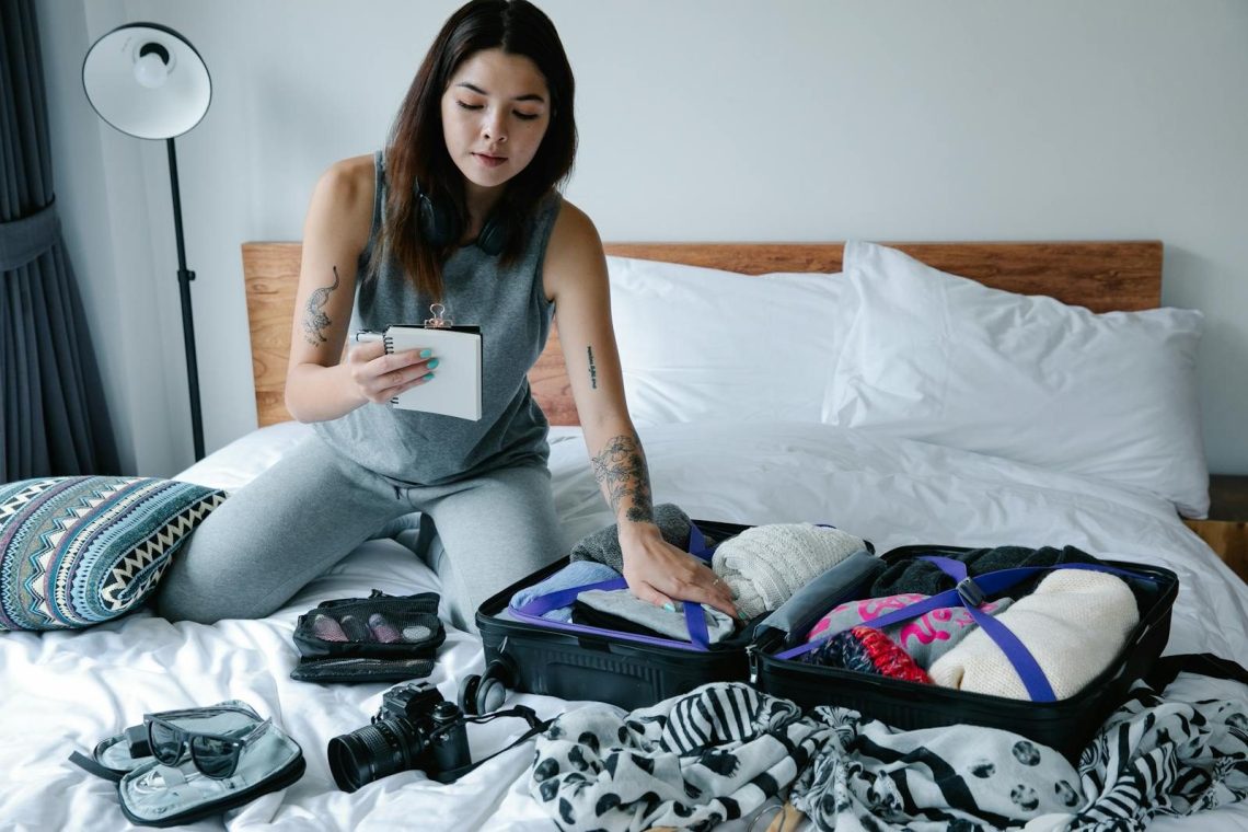 Woman sitting on bed packing a suitcase and taking notes for her travel plan.