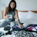 Woman sitting on bed packing a suitcase and taking notes for her travel plan.